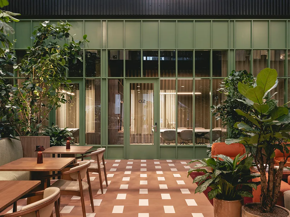 Green-framed glass meeting rooms viewed from the atrium with wooden tables and plants in the foreground