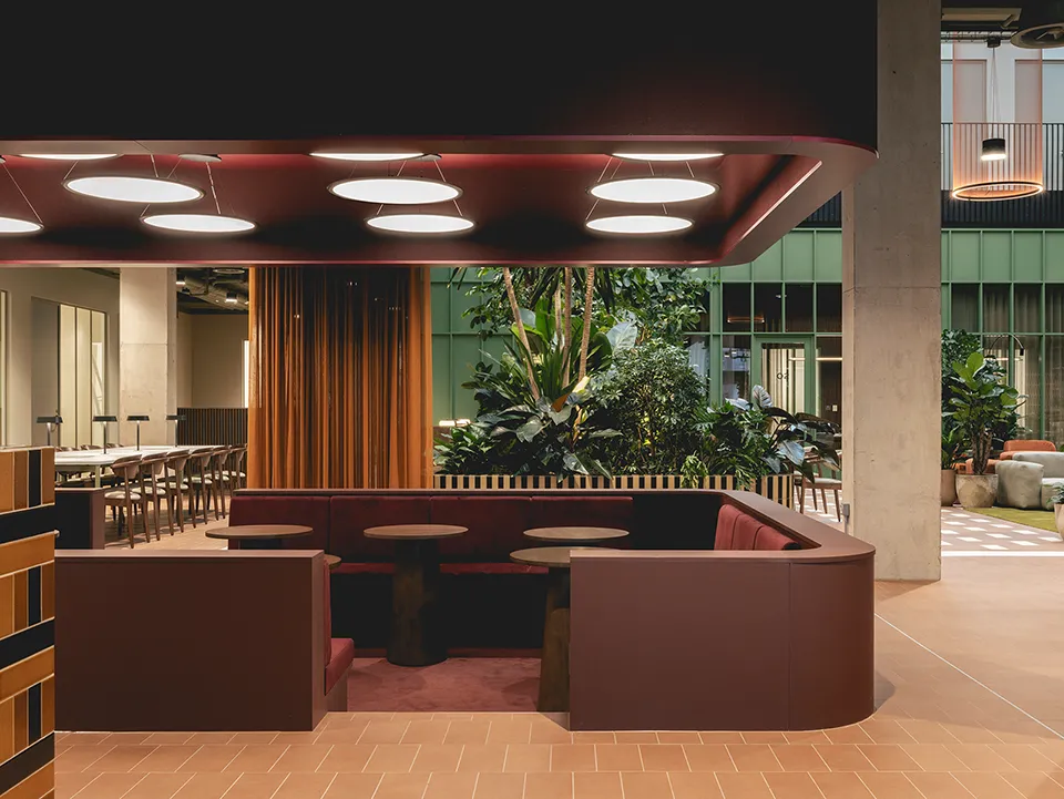 Curved burgundy booth seating beneath circular ceiling lights with atrium plants and amber curtains behind